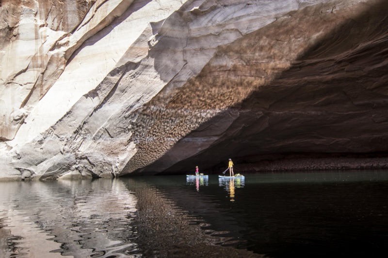 Two paddleboarders beneath a vast sandstone alcove on Lake Powell