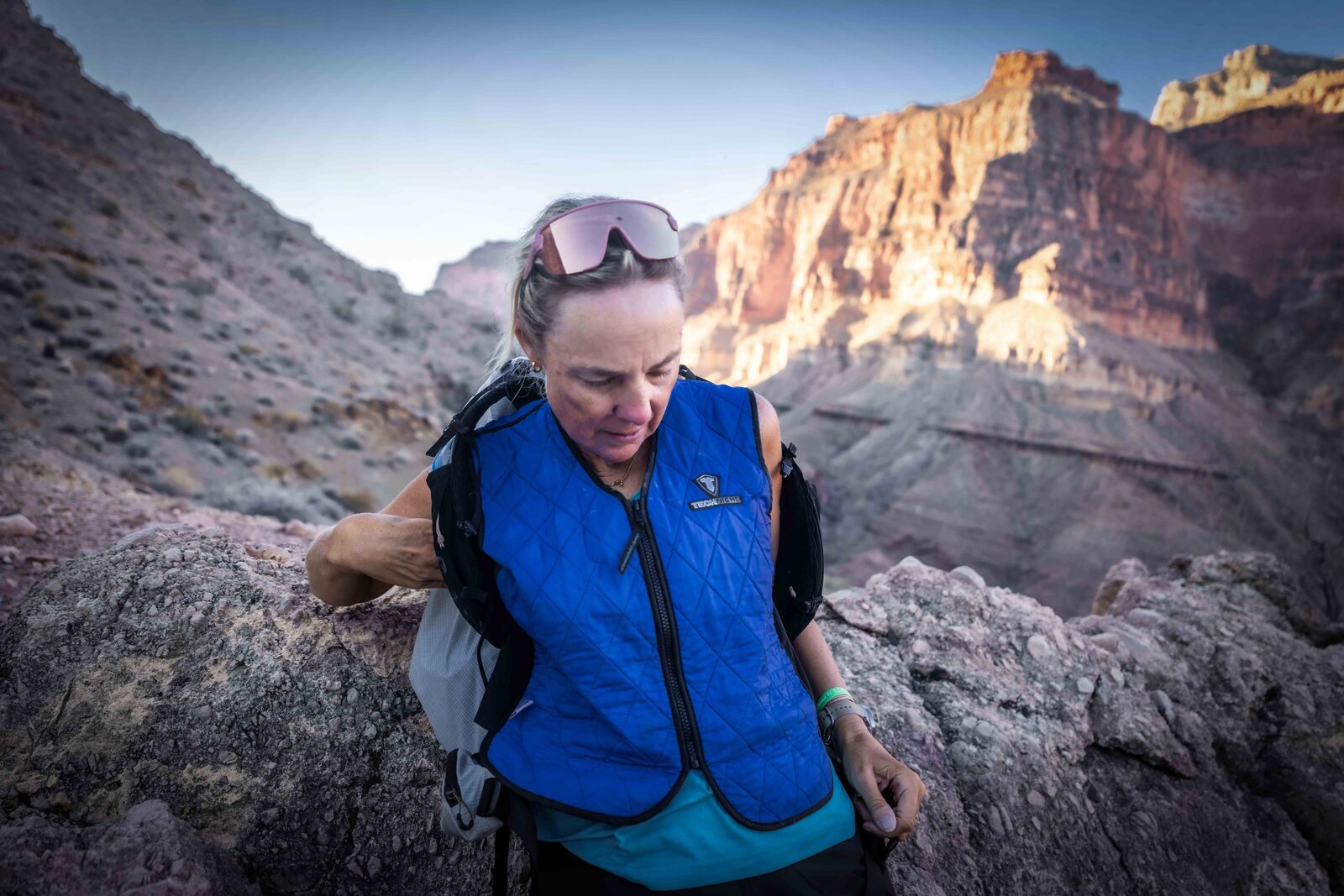 Mina Guli in a blue cooling vest on a rocky Grand Canyon ledge, early morning light on the canyon walls behind her