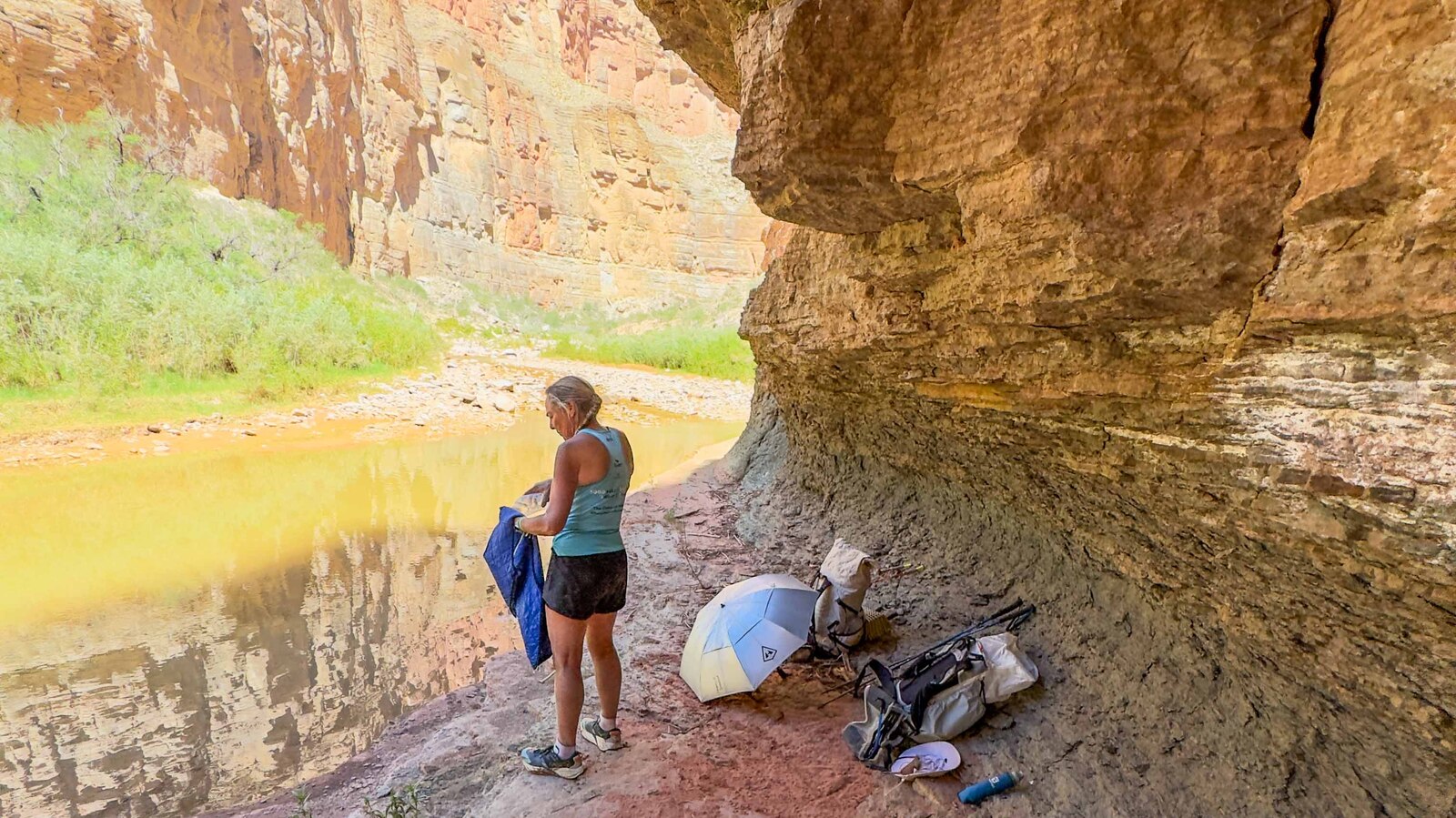 Sunny sheltering under a Grand Canyon overhang with sun umbrella and gear, golden river reflecting behind her