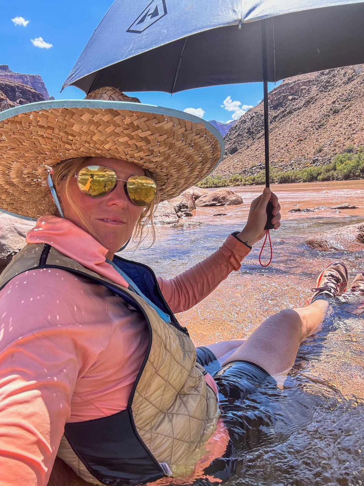 Sunny sitting in a Colorado River side channel, straw hat and sun umbrella deployed, feet in the water — heat management in the Canyon