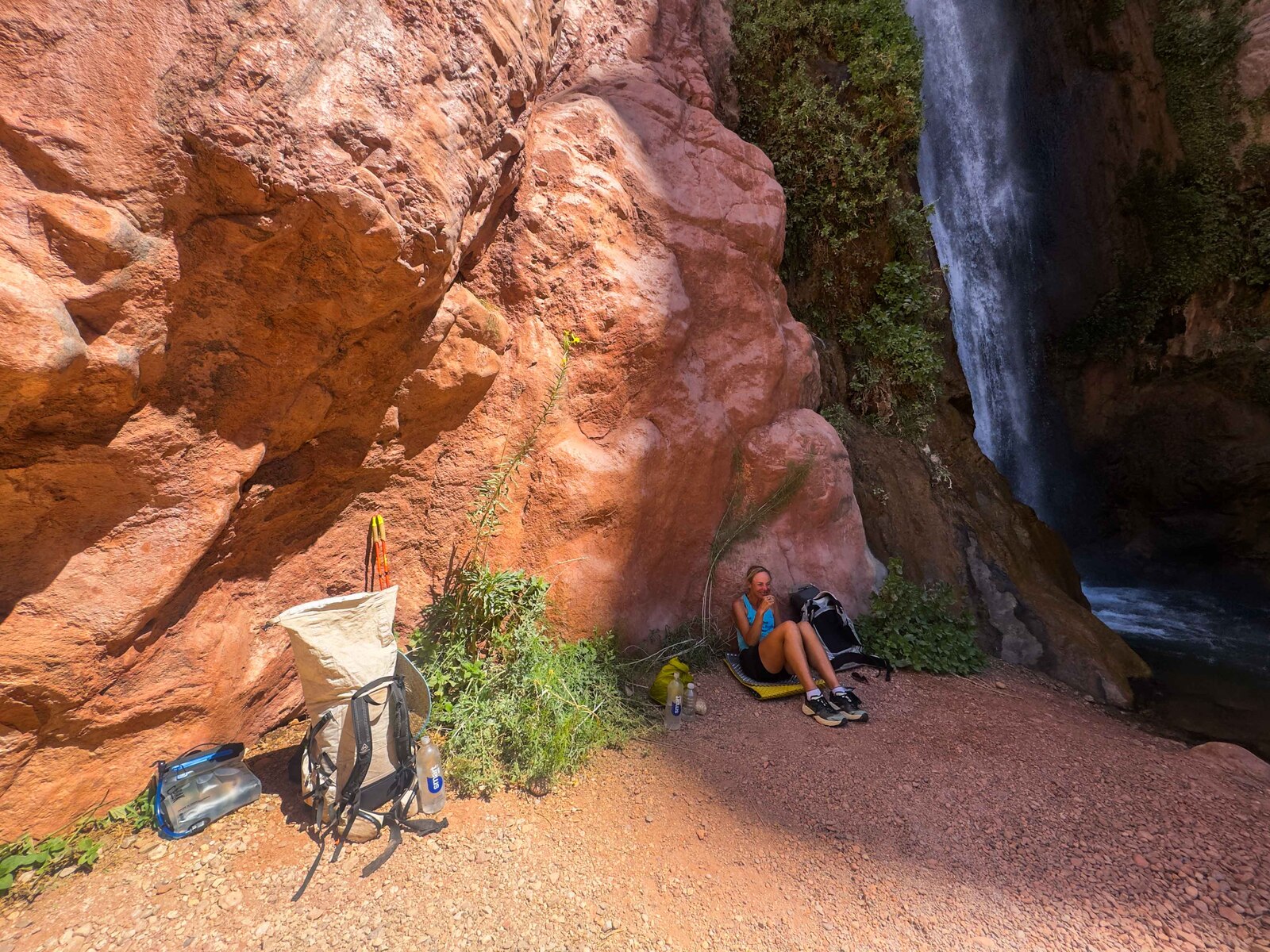 Sunny resting in the shade beside a waterfall in a red-rock Grand Canyon side canyon, gear and pack nearby