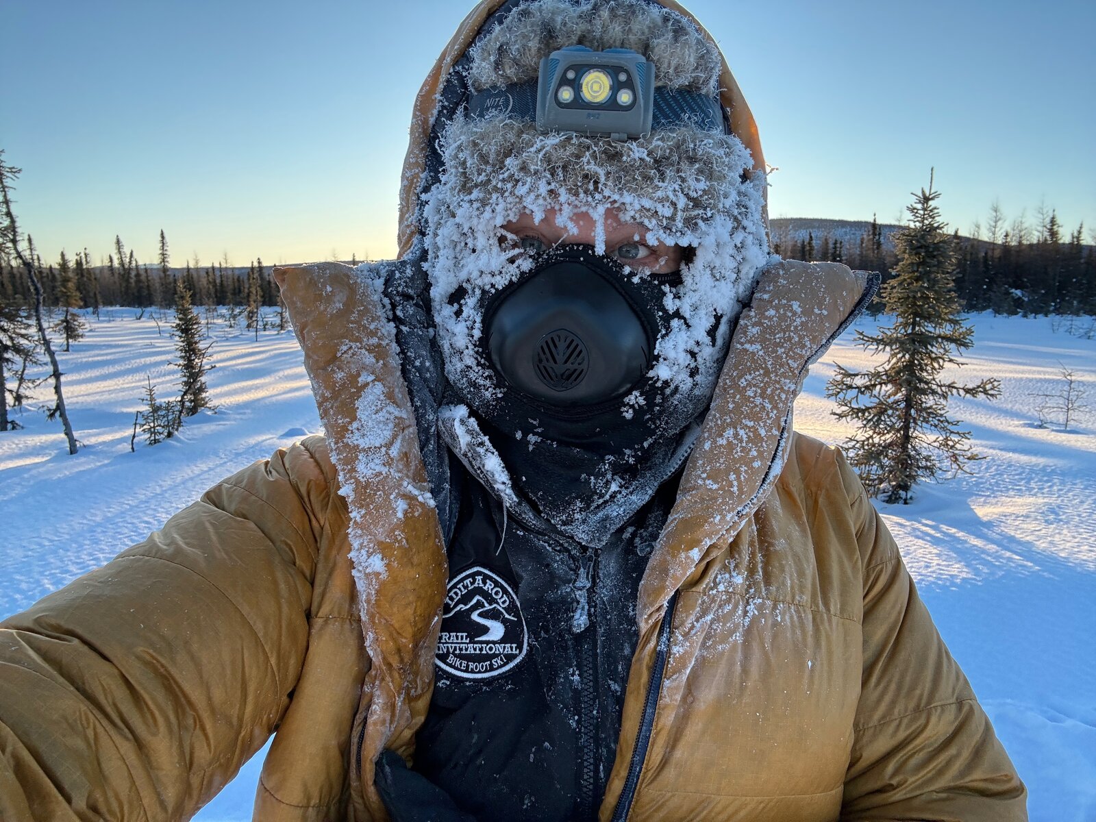 Sunny on the Iditarod Trail, face and balaclava completely covered in frost and ice, gold expedition parka, Alaskan taiga stretching behind her