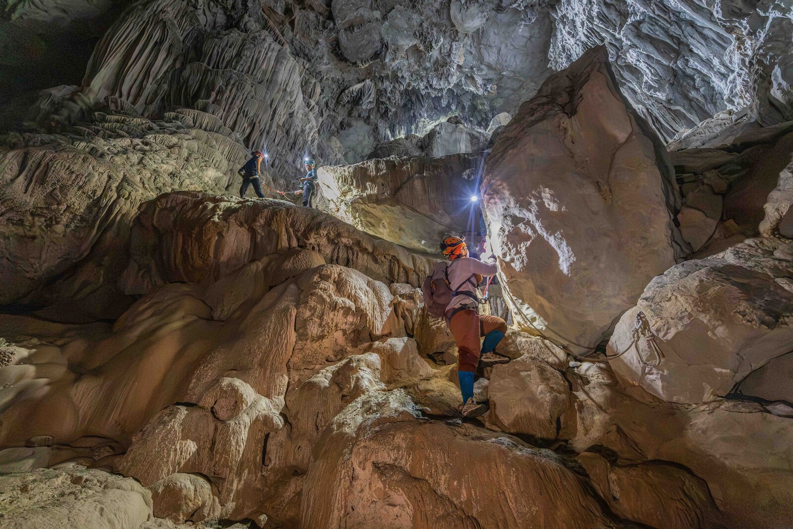 Multiple climbers pick their way across complex cave formations inside Son Doong, headlamps lighting the stone and stalactites above