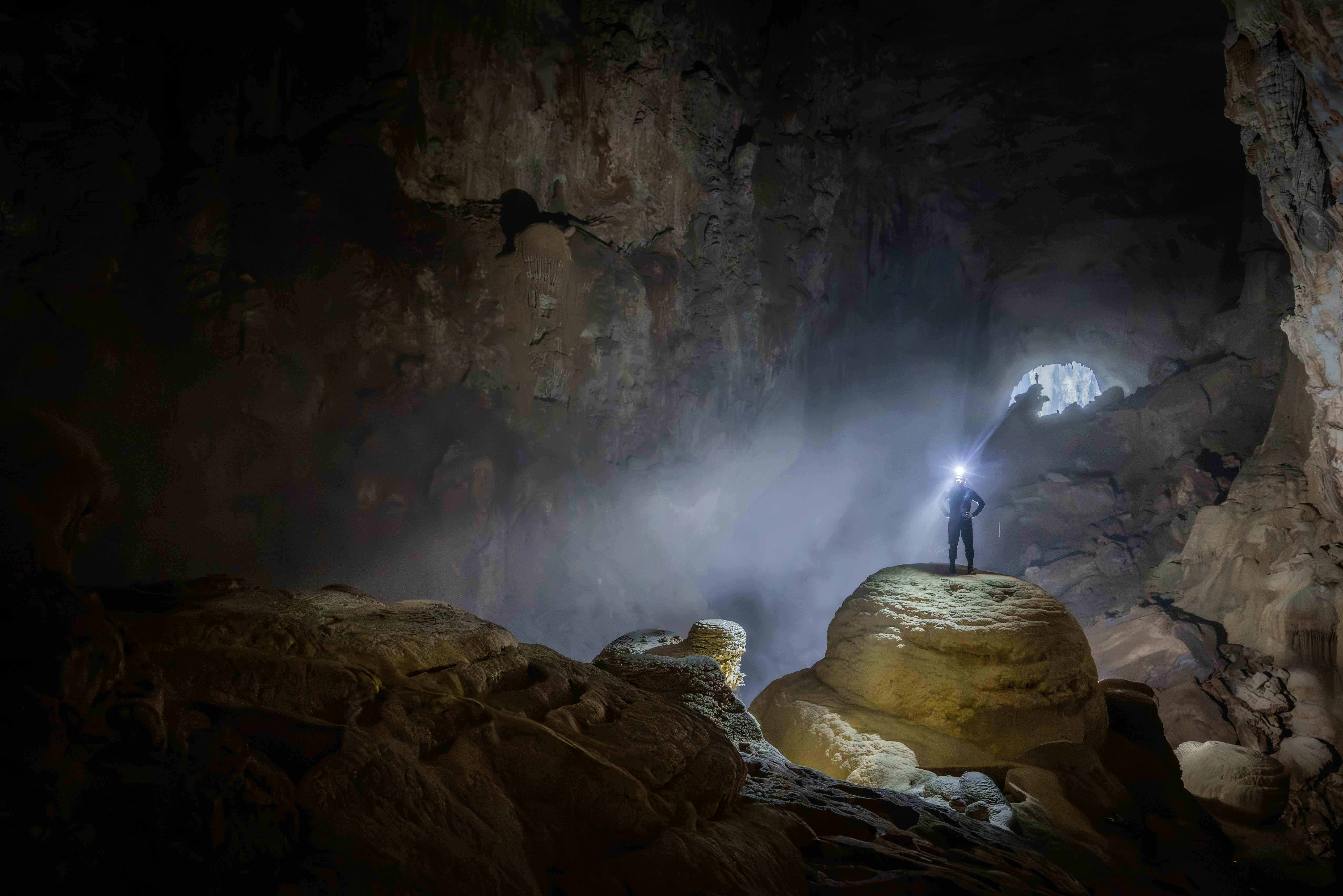 A lone figure stands on a cave formation inside Son Doong, the world's largest cave, a shaft of daylight cutting through the darkness above