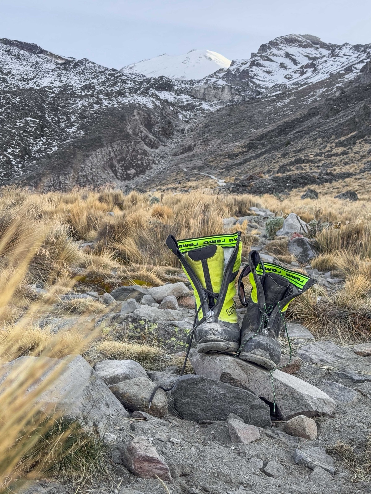 Two mismatched neon-green LOWA mountaineering boots perched on a rock in the foreground with the snow-capped summit of Pico de Orizaba rising in the background