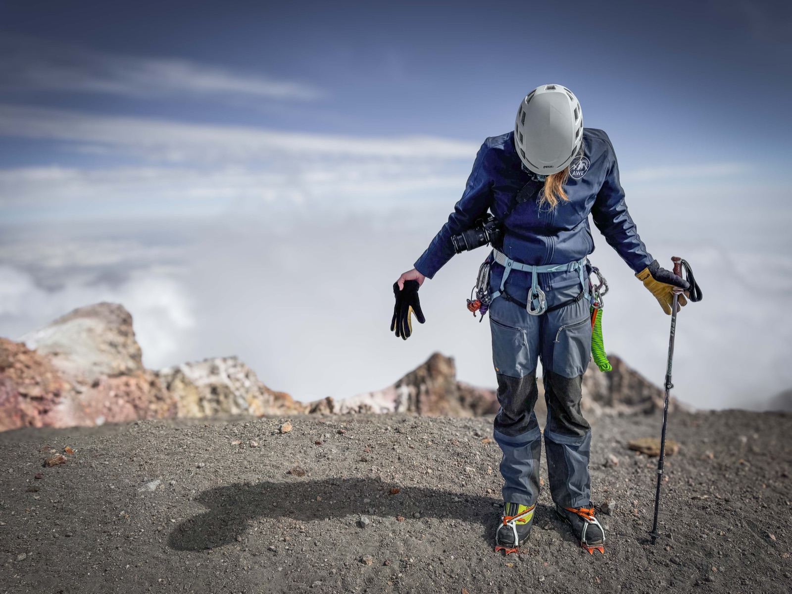 Sunny Stroeer standing on the gravel crater rim of Pico de Orizaba in helmet and harness, looking down at her crampon-clad boots, clouds far below