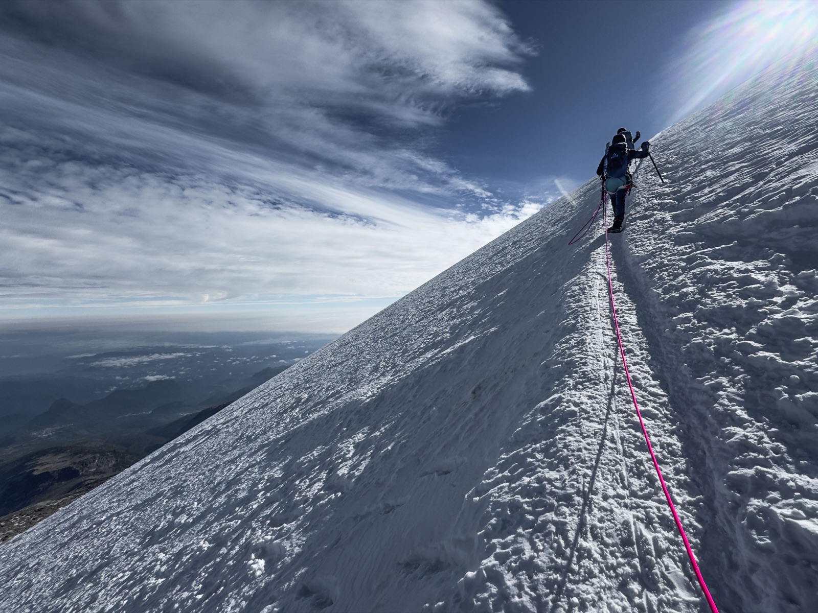 A rope team of two climbers ascending the steep snow slope of the Jamapa Glacier on Pico de Orizaba, pink rope trailing downhill, clouds and lowlands far below