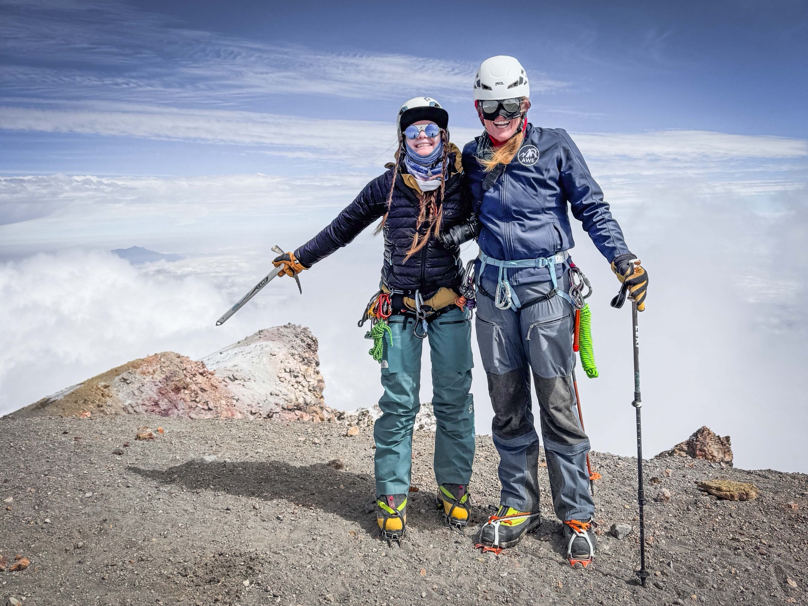 Two smiling climbers in helmets and harnesses standing arm in arm on the summit rim of Pico de Orizaba, one holding an ice axe, a sea of clouds behind them