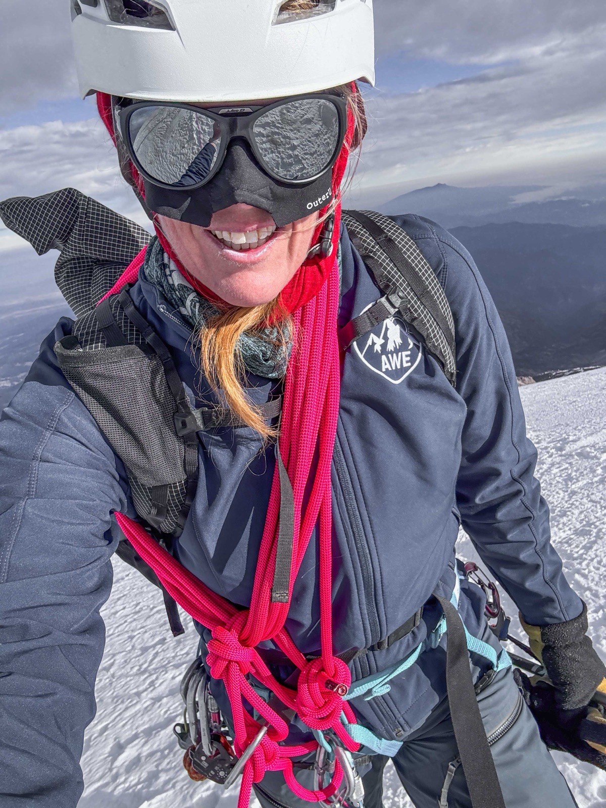 Summit selfie of Sunny Stroeer in a white helmet, glacier goggles, and navy AWE softshell with a coiled pink rope over her shoulders, snow and distant peaks below