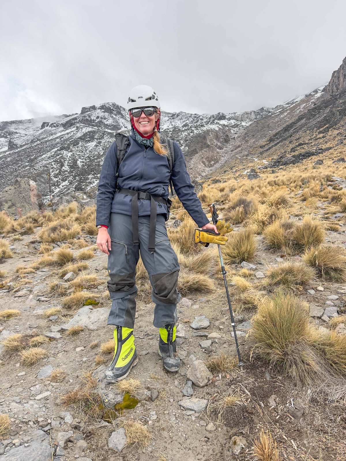 Sunny Stroeer smiling in helmet and softshell among golden tussock grass below the snow-dusted Labyrinth on Pico de Orizaba, neon LOWA boots on her feet