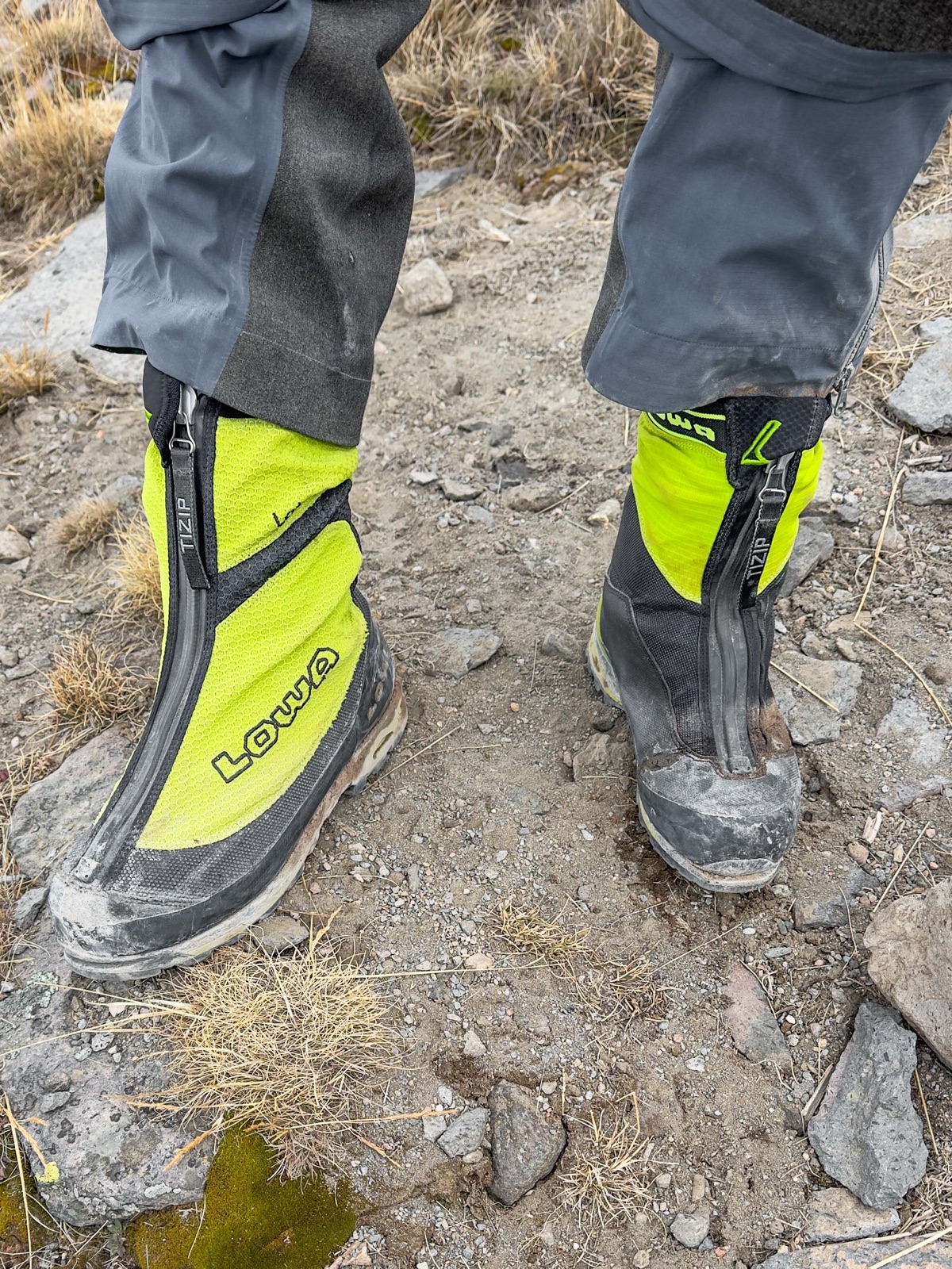 Looking down at two neon-green-and-black LOWA mountaineering boots being worn on a rocky trail - both left boots, one visibly larger than the other
