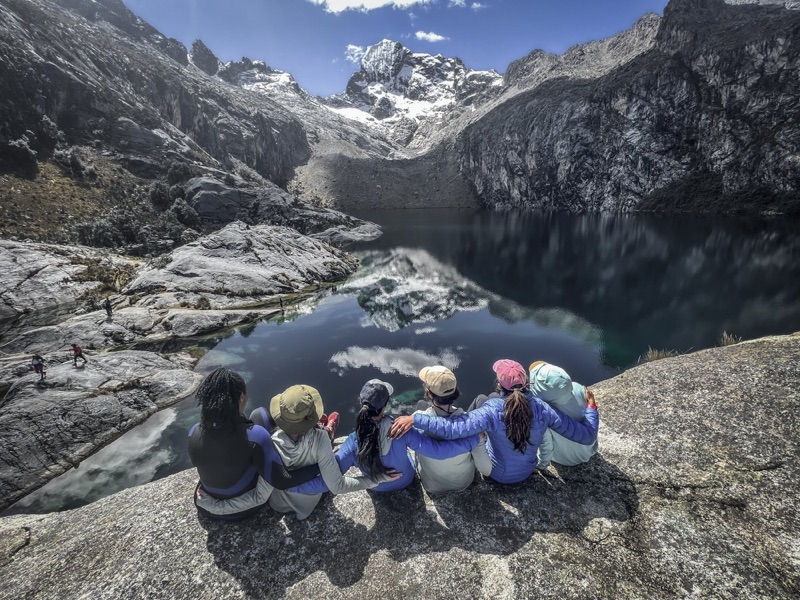 Five women sitting arm in arm above a dark alpine lake