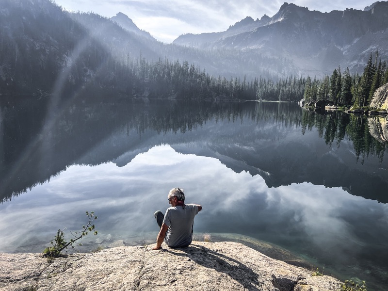 Man sitting at the edge of a still mountain lake at dawn