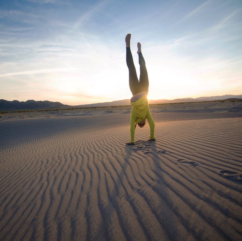 A person in a yellow top holds a handstand on rippled sand dunes at sunset with desert mountains on the horizon