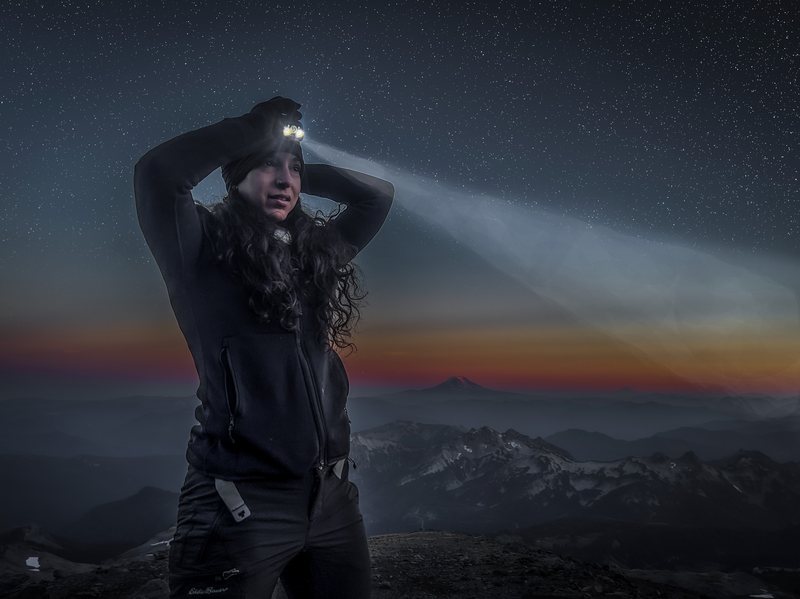 A woman wearing a lit headlamp stands on a high ridge at dusk with stars overhead and a distant snow-capped volcano behind her