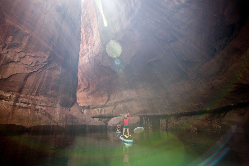 Two paddleboarders cross a flooded slot canyon as sunbeams angle down the sandstone walls