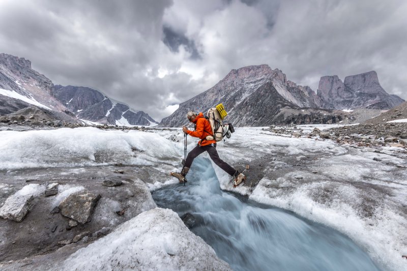 A backpacker in an orange jacket leaps across a meltwater stream on a glacier beneath rocky peaks and heavy clouds