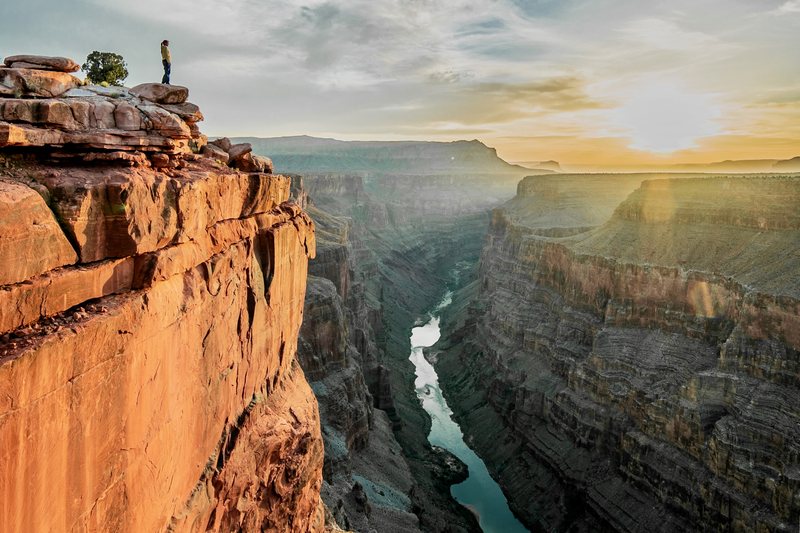 A tiny figure stands on a cliff edge above the river in the inner gorge of the Grand Canyon at sunrise
