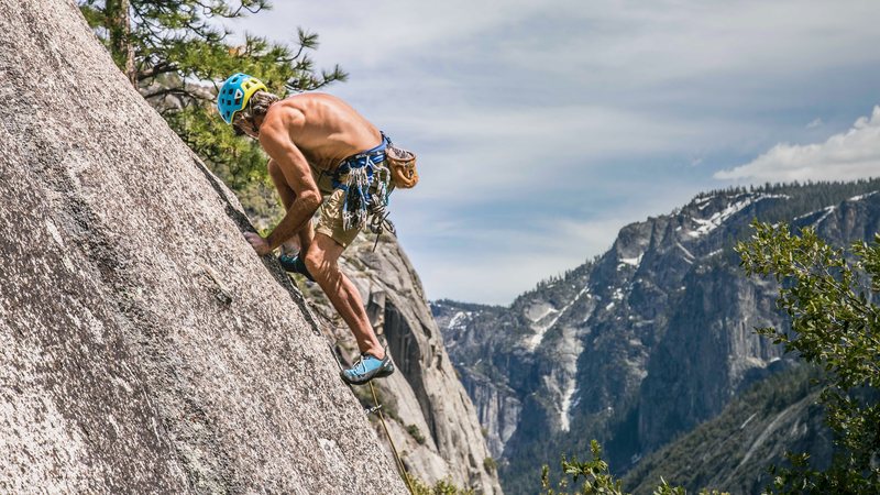 A helmeted climber moves up a granite slab high above a forested glacial valley