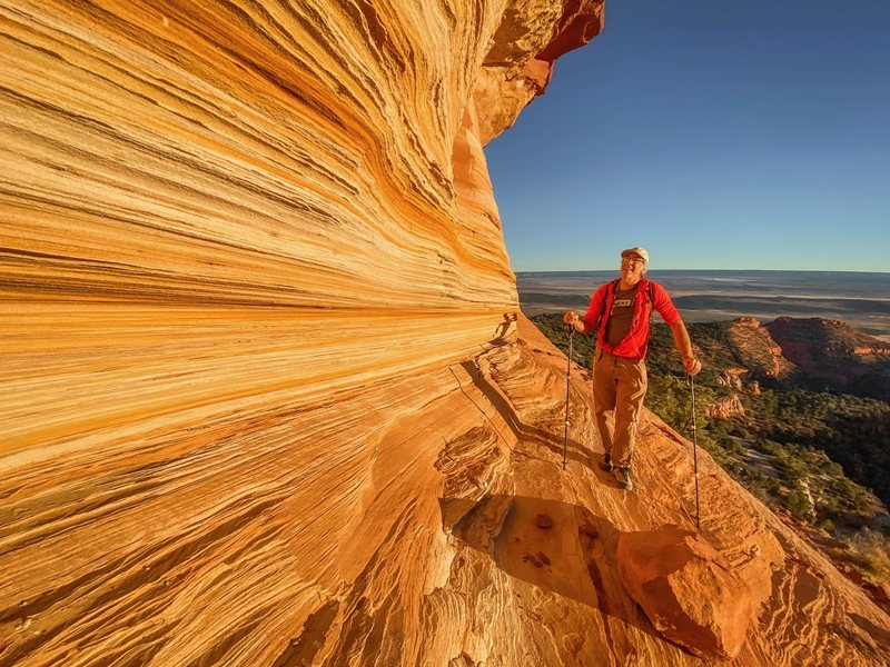 A hiker with trekking poles stands on a narrow ledge beneath a striped sandstone overhang above a desert valley