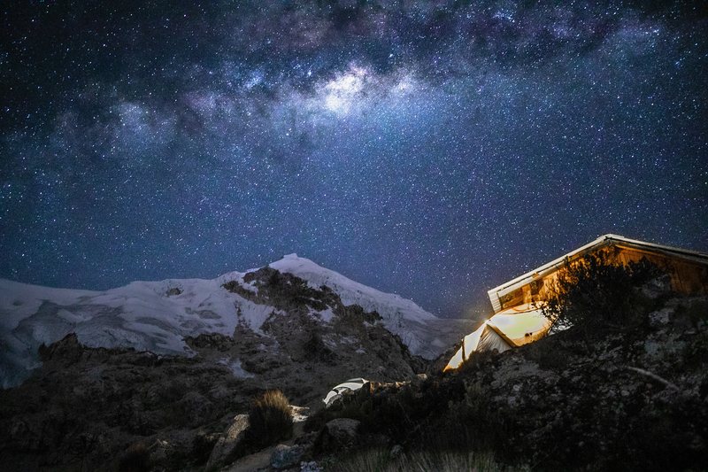 The Milky Way arcs over a snow-covered peak and a small mountain hut with glowing tents below