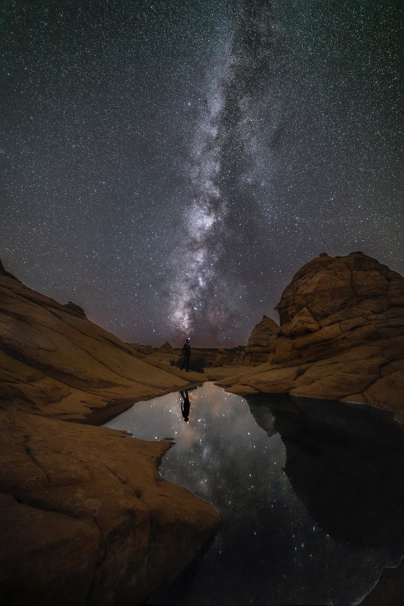 The Milky Way rises between sandstone domes and is mirrored in a desert pool where a tiny figure stands