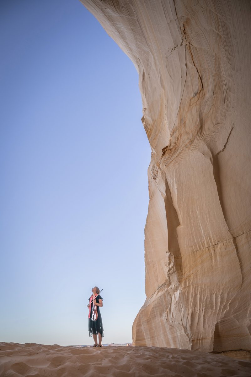 A slightly motion-blurred figure stands on sand at the base of a towering pale sandstone wall