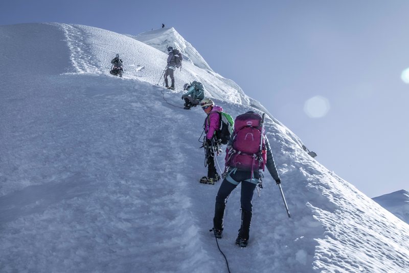 Mountaineers with packs and ice axes ascend a steep snow slope toward climbers higher on the ridge