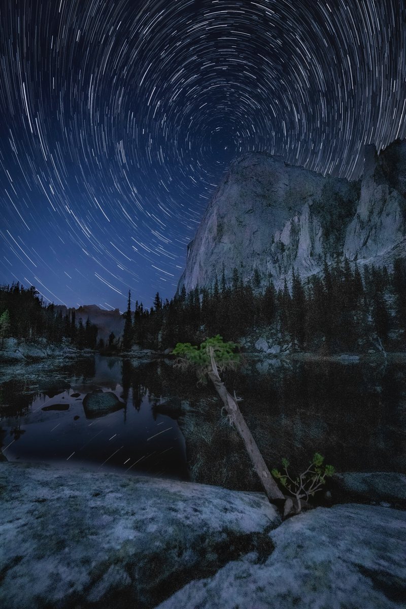 Circular star trails spin above a granite cliff and a calm lake with a fallen log in the foreground