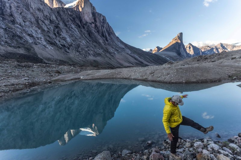 A hiker in a yellow jacket balances on rocks beside a still tarn that reflects a sheer granite peak