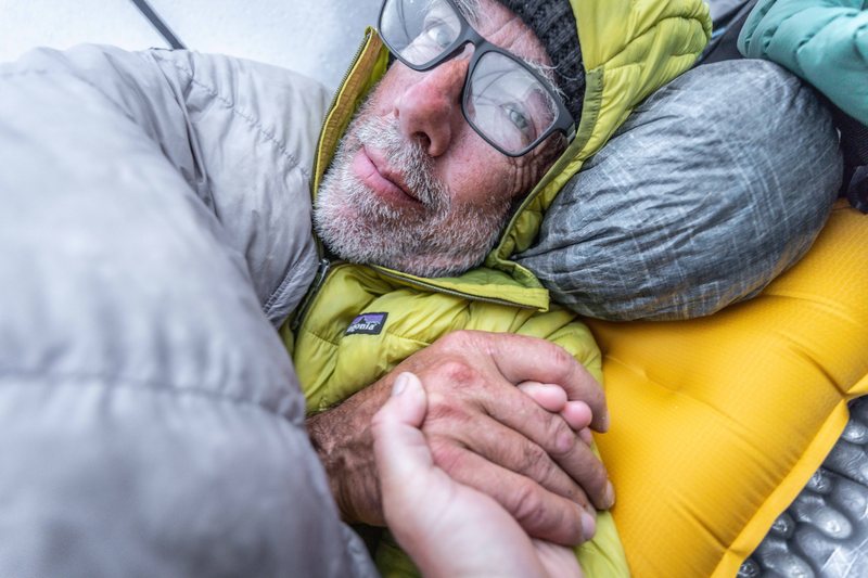 A bearded climber in glasses and a yellow puffy jacket lies in a sleeping bag inside a tent, holding the photographer's hand