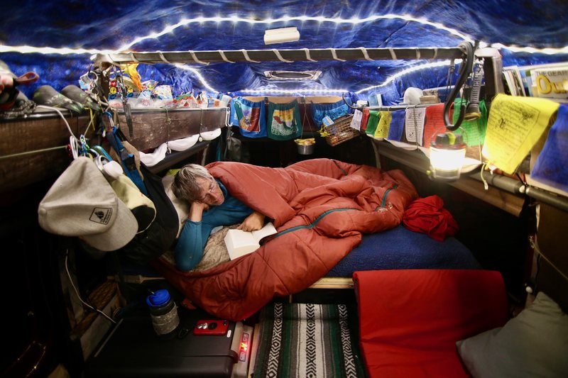 A person reads a book in bed inside a camper van decorated with string lights and prayer flags